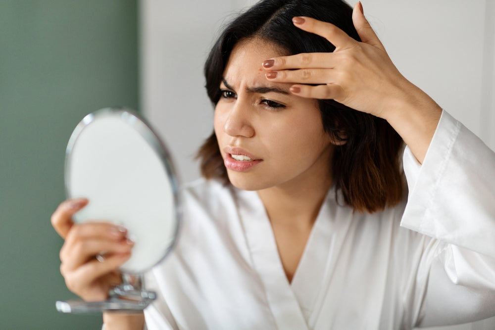Image of a brunette woman holding up a magnifying mirror and pulling at the dull skin on her forehead.