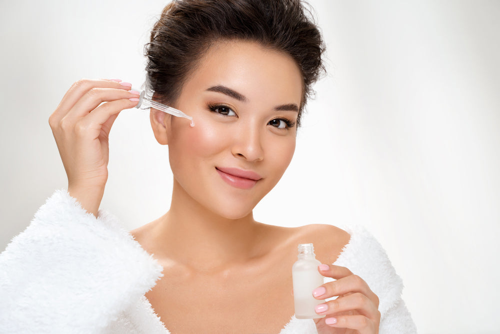 Young woman in a white robe using a dropper to apply serum to her face.