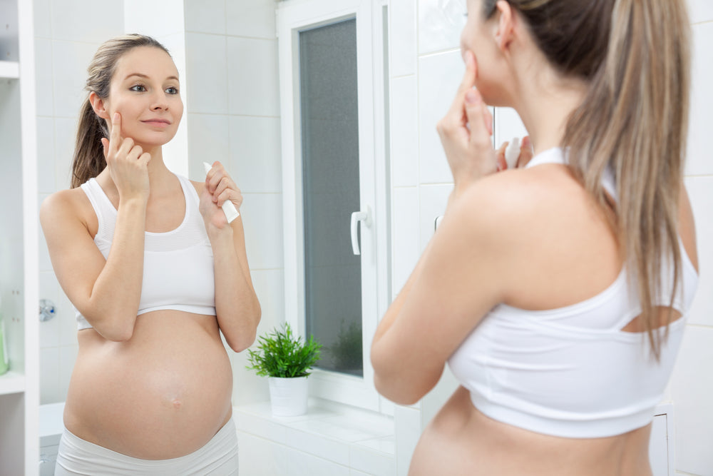Pregnant blonde woman putting on face cream while looking at herself in the bathroom mirror.