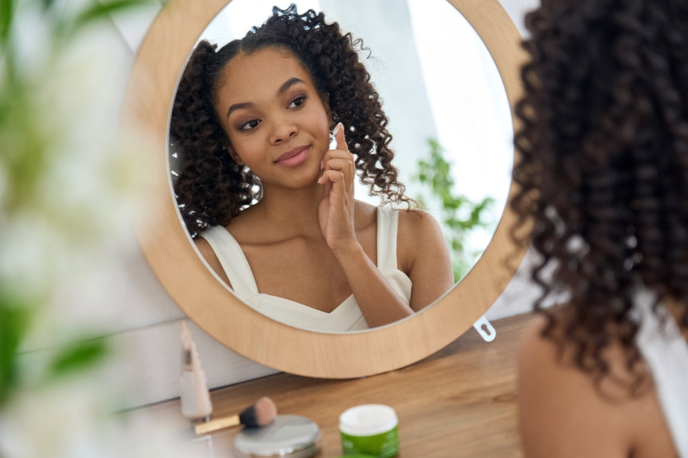 Black teen girl applying cream to her face while sitting at a wood vanity.