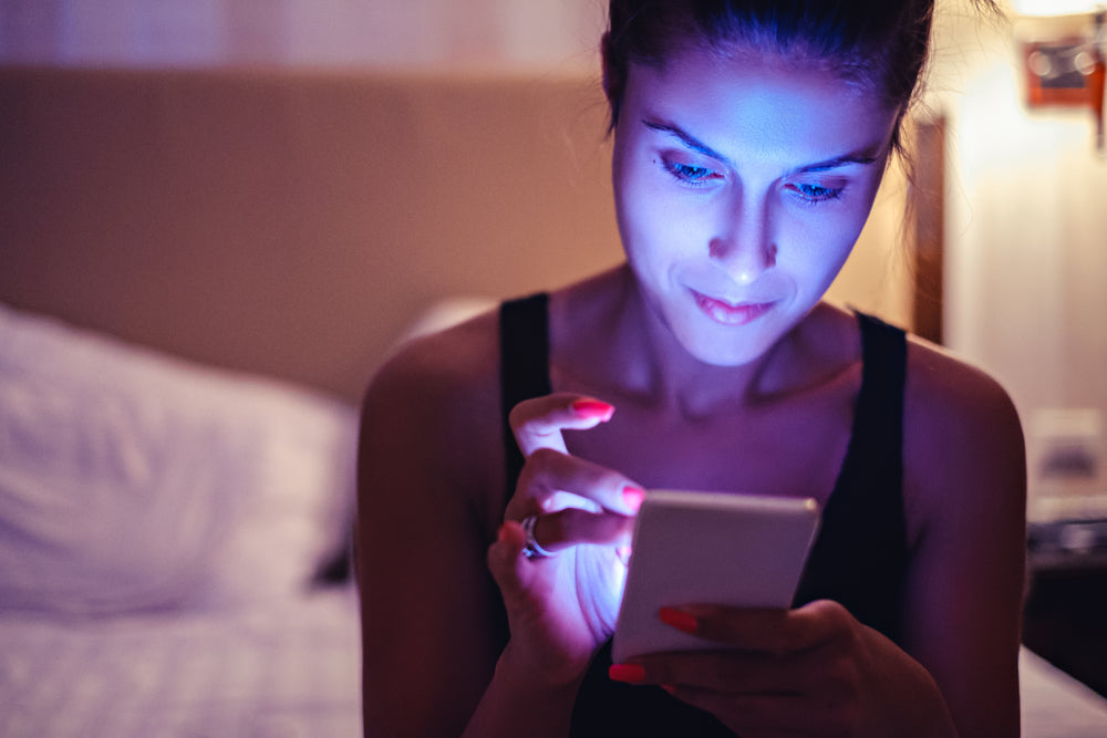 Woman in dark room sitting on bed looking at a device with strong blue light emissions