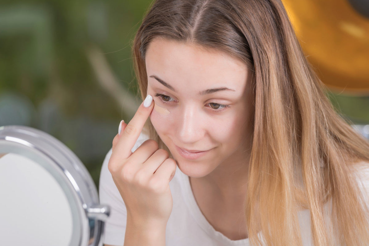 Brunette woman leaning in front of a magnifying mirror and applying primer under her eye with one finger.