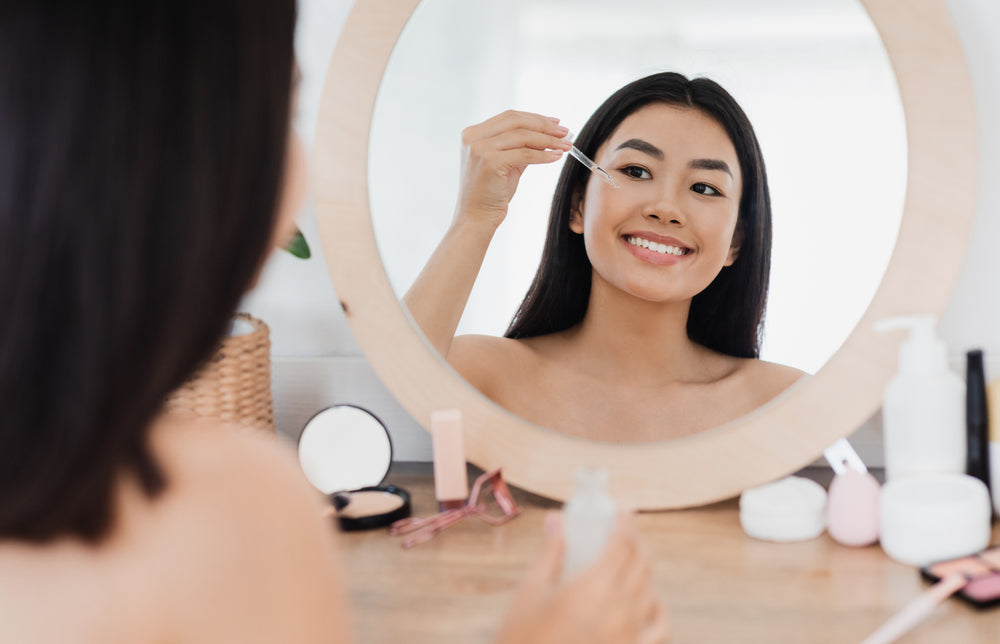 Asian woman sitting at a vanity with various products in front of her and applying serum with a dropper to her cheek.