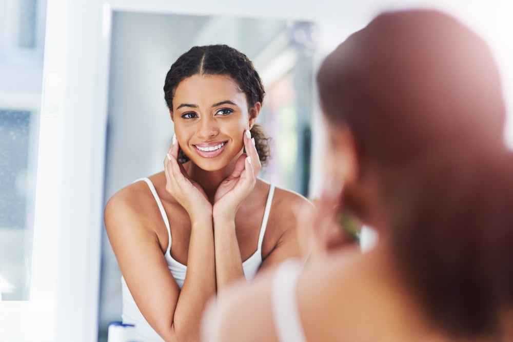 Young Black woman looking at her skin in the mirror.