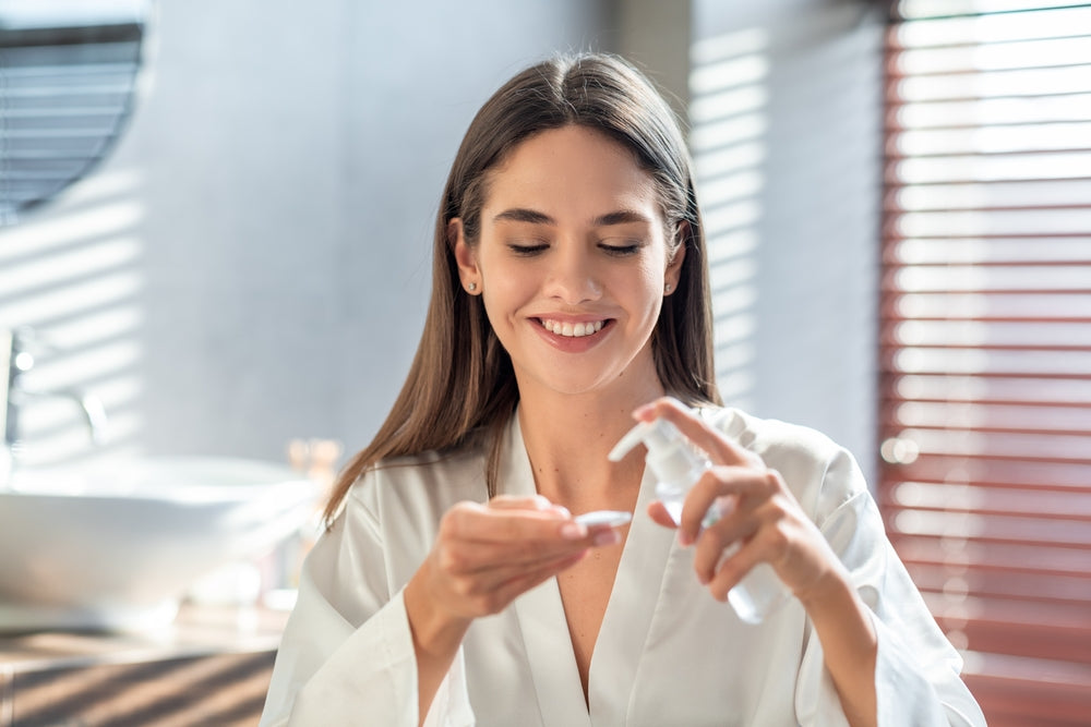 Young woman standing in the bathroom applying a skin product to a cotton round.