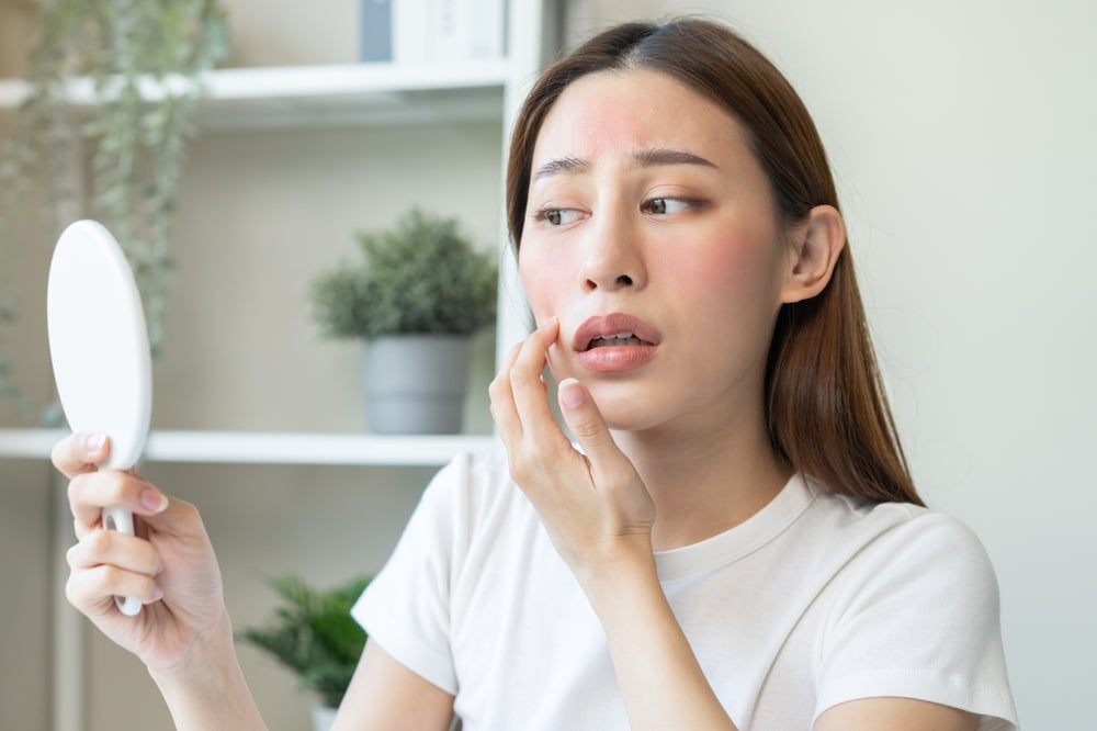 Young asian woman holding up a white mirror and touching her sunburned cheek with a worried expression on her face.