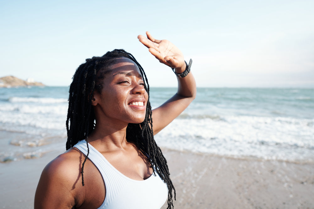 Young Black woman standing on a beach and holding her hand up to block the sun from her eyes.