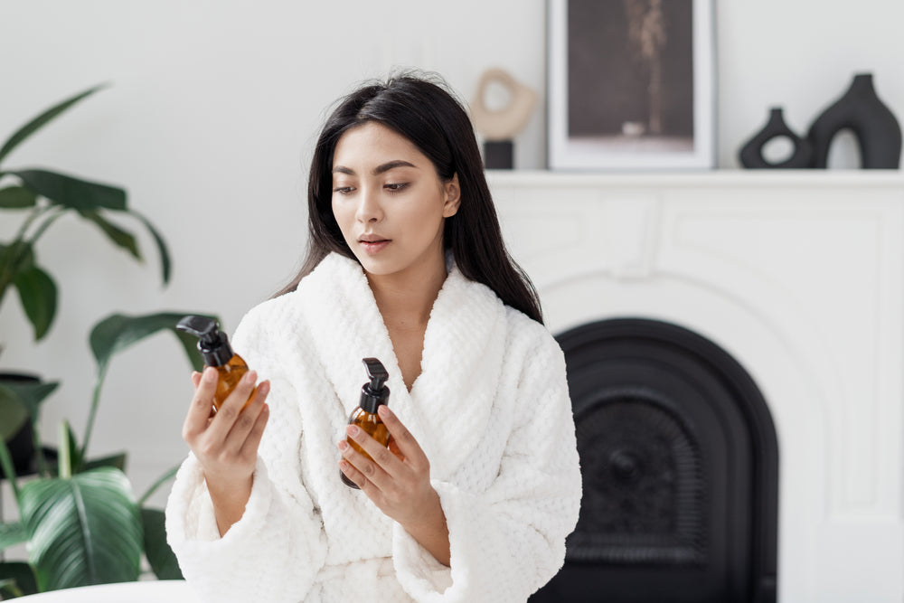 Young woman looking at the active ingredients on the labels of two skincare products while sitting in her living room wearing a robe.