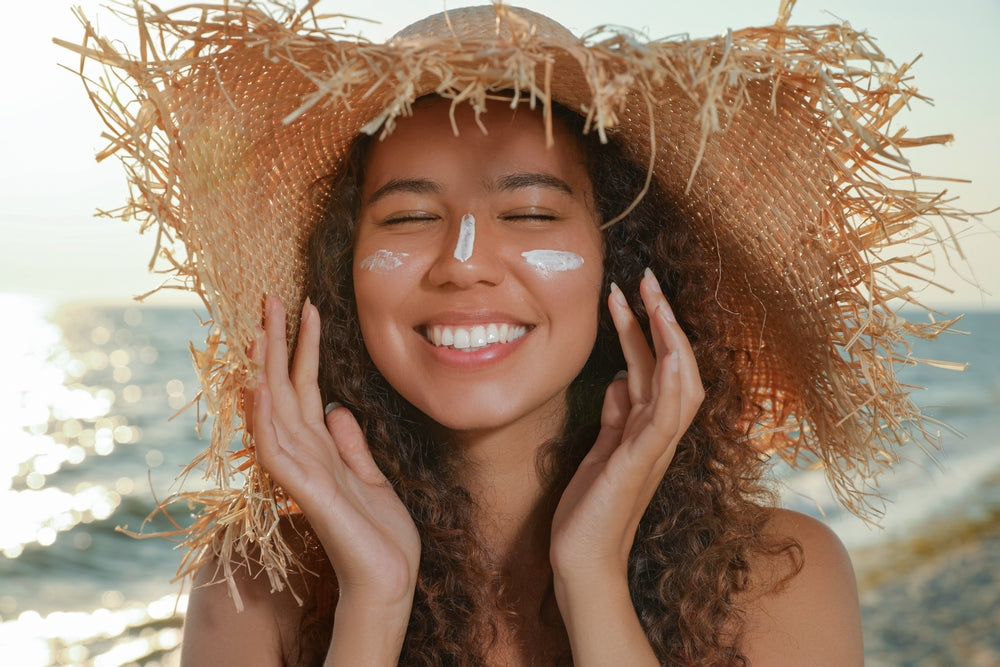 Smiling woman in straw hat at the beach applying high SPF sunscreen to her face.