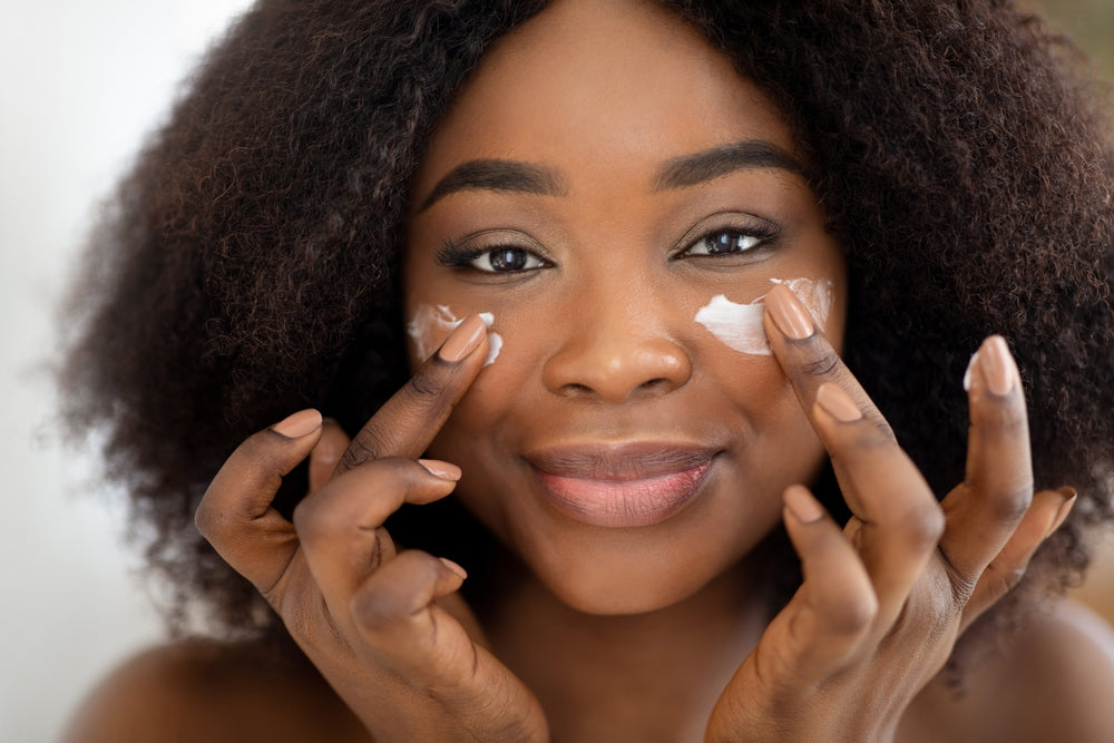 Image of a young Black woman applying cream under her eyes.